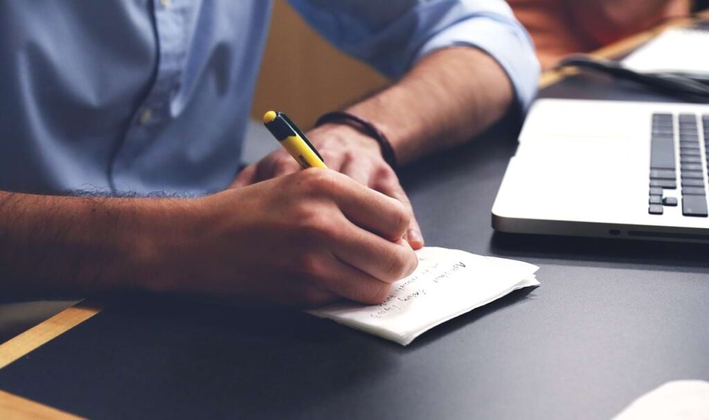 Close-up of a professional writing notes beside a laptop, symbolizing internal reflection after comparing LinkedIn profiles